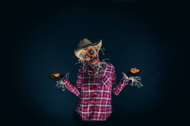 A creepy scarecrow with a pumpkin face holding two little pumpkins.