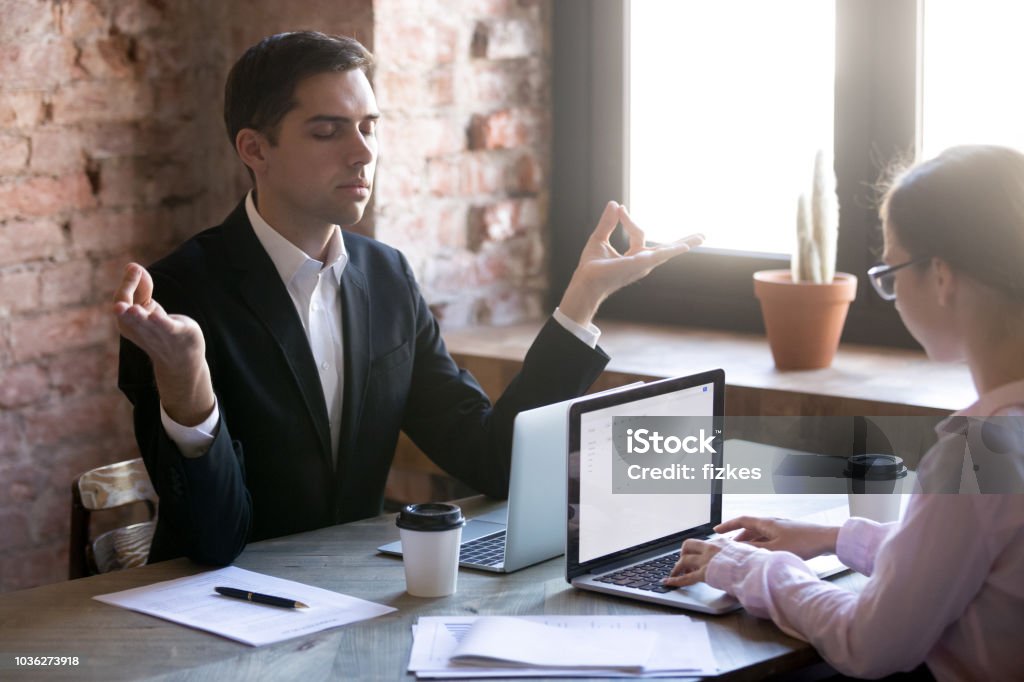 Meditating at workplace Male worker sitting in workplace in front of laptop and taking break for meditation, woman colleague. Thinking, focusing, stress relief, healthy good habits, mental health, mindful lifestyle concepts Adult Stock Photo