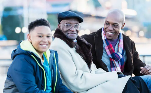 A multi-generation African-American family spending time together, relaxing on a city waterfront. The senior man in the middle is sitting between his grandson and his great grandson. The great grandfather is 79 years old. They are all smiling and relaxed, looking at the camera.