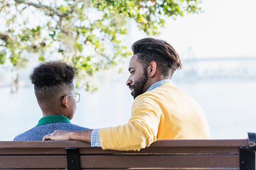 Rear view of a mixed race African-American and Hispanic father and his teenage 13 year old son. They are sitting outdoors on a park bench with a view of a city waterfront. The man is giving the teenage boy some fatherly advice.