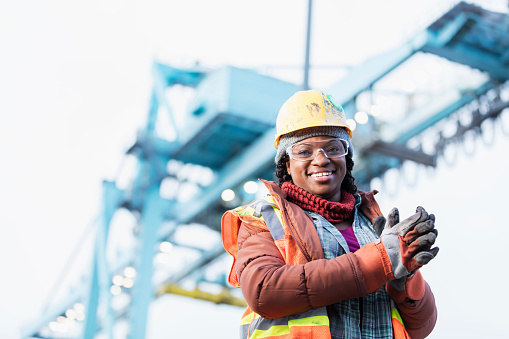 A mid adult African-American woman in her 30s wearing a hard hat, safety vest and safety goggles, a dock worker working at a shipping port. A gantry crane is out of focus in the background.
