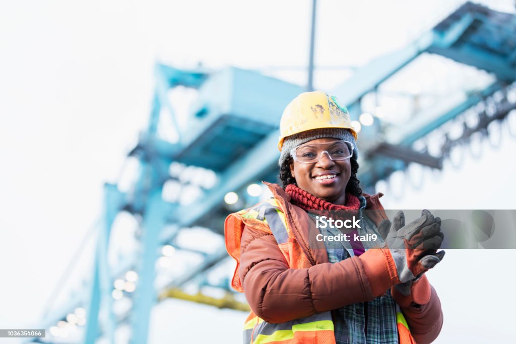 African-American woman working at shipping port A mid adult African-American woman in her 30s wearing a hard hat, safety vest and safety goggles, a dock worker working at a shipping port. A gantry crane is out of focus in the background. Construction Worker Stock Photo