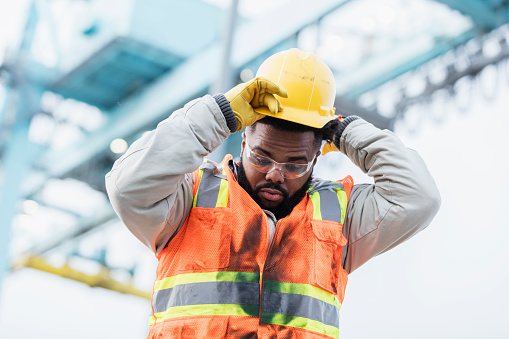 A young African-American mam in his 20s working at a shipping port, wearing a safety vest and safety glasses, putting on a hardhat. A gantry crane is out of focus in the background.