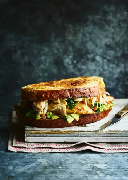 A chicken and avocado sandwich on toasted bread sat on a wooden board and napkin with a knife on the side.