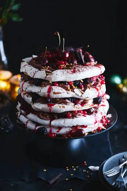 A five layer cherry chocolate pavlova with red currants, blackberries, cherries, chocolate curls, and golden sprinkles and stars with Christmas baubles and lights in the background