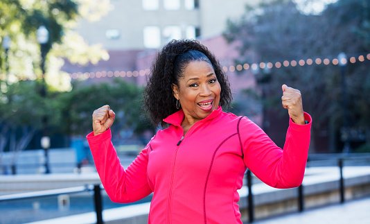A senior African-American woman in her 60s wearing a pink sweatshirt, exercising in the city. She is smiling a the camera, flexing her biceps, showing her strength.