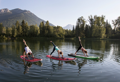 Three people practicing yoga on a paddle board with mountains in the background.