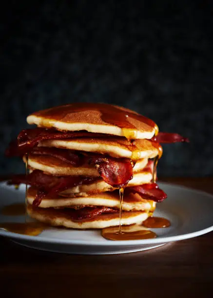 A stack of pancakes from the side, with bacon and maple syrup.