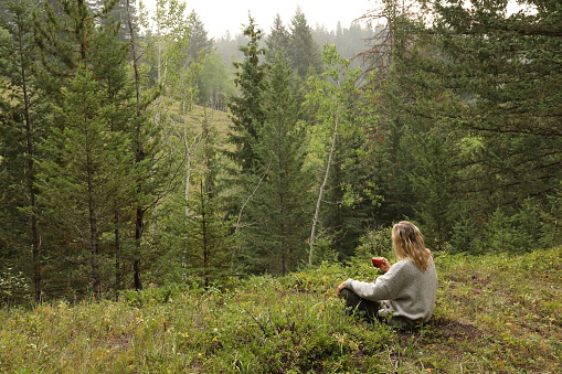 Woman drinks morning coffee, while sitting in forest meadow