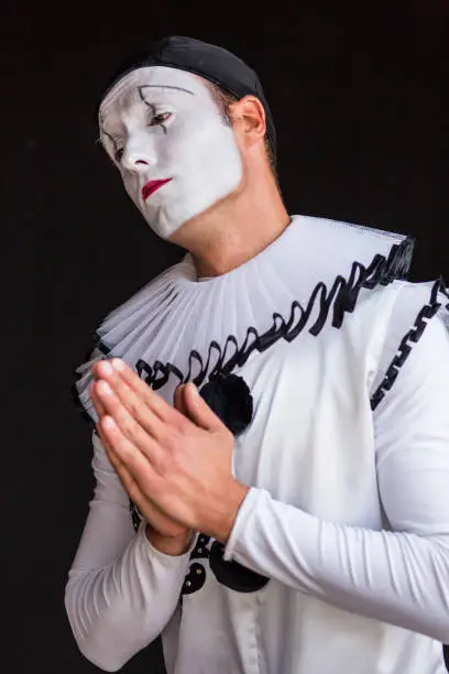 Beautiful close up photography of a mime artist, holding his hands for praying and his head cast aside with a sad facial expression. The portrait is on a black background. The clown is dressed in white costume with big black buttons and a black hat. His face is white, painted with make up. The actor has brown eyes. The mimic artist is mimicking emotions. The model is a professional mime artist from Ukraine. He is working in a circus, doing his show as a solo act. Mime artist is making facial expressions, looking directly into camera. Mummer or pantomime are other words describing this studio shot.