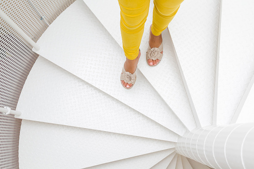 Young woman's legs in yellow trousers and sandals going down on white spiral stairs. Top view.