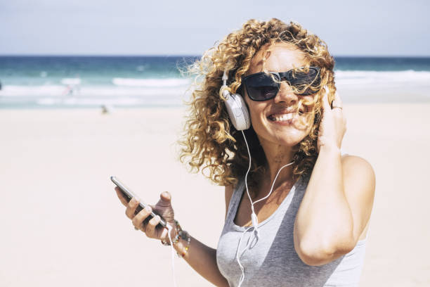 joyeux attrayant beau caucasien moyen age femme souriant à la plage dans un lieu tropical tout en ecoute la musique avec le téléphone intelligent. personnes bénéficiant des vacances et la liberté de travail vacances - tourist resort audio photos et images de collection