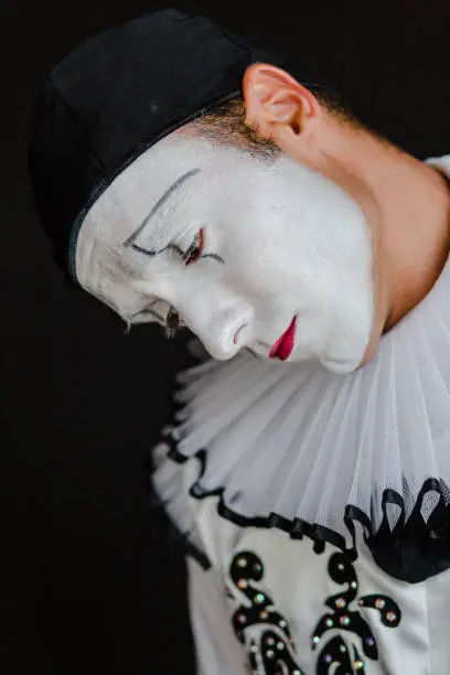 Beautiful close up photography of a mime artist, looks like hanged. The portrait is on a black background. The clown is dressed in white costume with big black buttons and a black hat. His face is white, painted with make up. The actor has brown eyes. The mimic artist is mimicking emotions. The model is a professional mime artist from Ukraine. He is working in a circus, doing his show as a solo act.