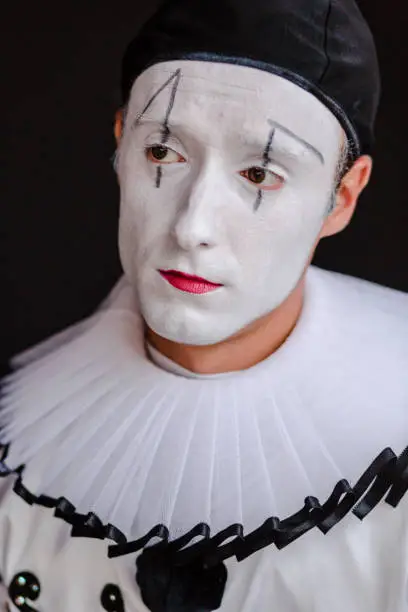Beautiful close up photography of a mime artist. looking away from the camera. The portrait is on a black background. The clown is dressed in white costume with big black buttons and a black hat. His face is white, painted with make up. The actor has brown eyes. The mimic artist is mimicking emotions. The model is a professional mime artist from Ukraine. He is working in a circus, doing his show as a solo act.