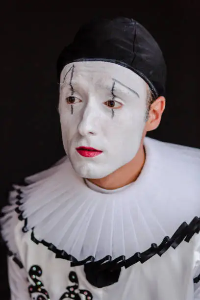 Beautiful close up photography of a mime artist. looking away from the camera. The portrait is on a black background. The clown is dressed in white costume with big black buttons and a black hat. His face is white, painted with make up. The actor has brown eyes. The mimic artist is mimicking emotions. The model is a professional mime artist from Ukraine. He is working in a circus, doing his show as a solo act.