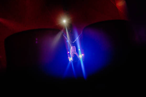 Photography of a real acrobat performing in a circus play, balancing, jumping, and standing on his hands. The circus performer is in the spotlight. The acrobat is well balanced and brave. The gymnast is dressed in blue pants and white shirt. He is in a live act. The light show is spectacular. The background is dark and there is light cast on the performer. It is a dangerous act. The circus acrobat is trained and capable.
