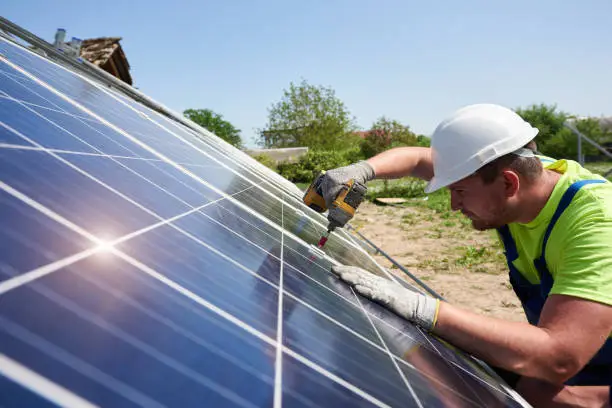 Profile of professional technician working with screwdriver connecting shiny solar photo voltaic panel to metal platform system on summer rural landscape and blue sky copy space background.