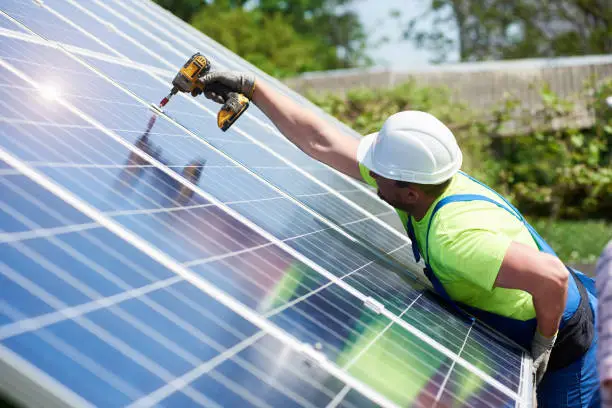 Professional technician working with screwdriver connecting solar photo voltaic panel to metal platform on summer rural landscape background. Stand-alone exterior solar panel system installation.