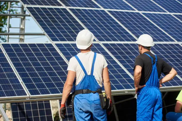 Back view of two workers in blue overalls standing in front of high exterior solar panel photo voltaic system installed on steel platform. Renewable ecological green energy production concept.