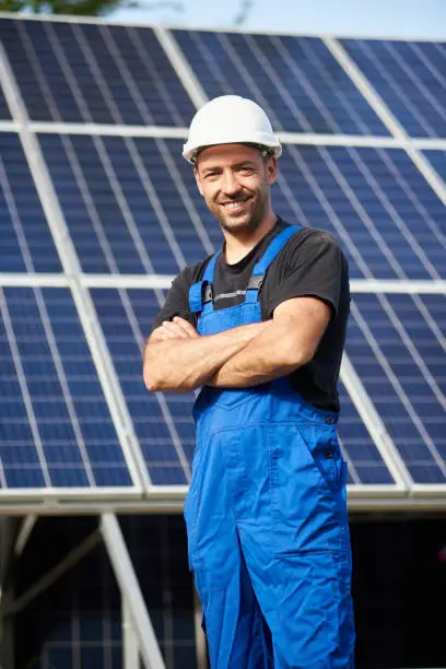 Portrait of smiling successful male engineer technician standing in front of unfinished high exterior solar panel photo voltaic system blue shiny surface.