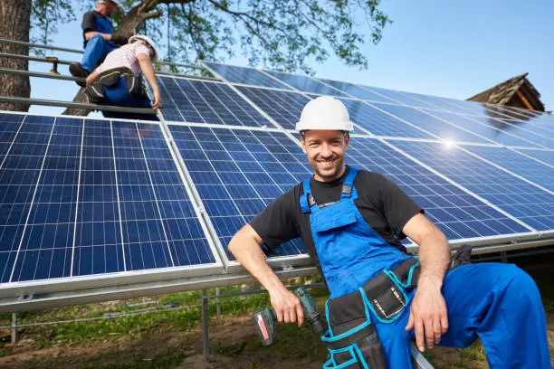 Portrait of happy technician with electrical screwdriver in front of unfinished high exterior solar panel photo voltaic system with team of workers on high platform.