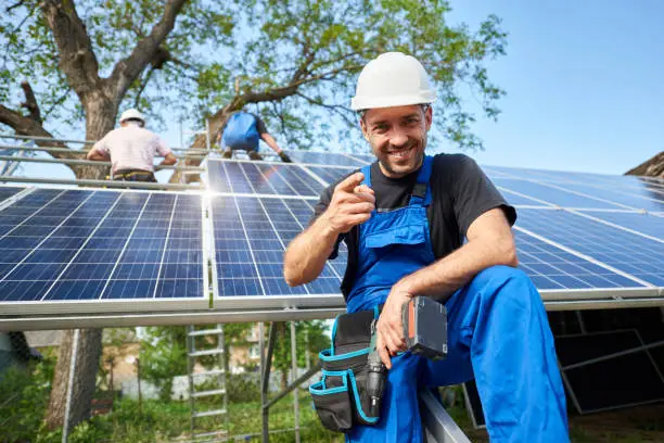 Portrait of smiling technician with electrical screwdriver pointing to camera in front of unfinished high exterior solar panel photo voltaic system with team of workers on high platform.