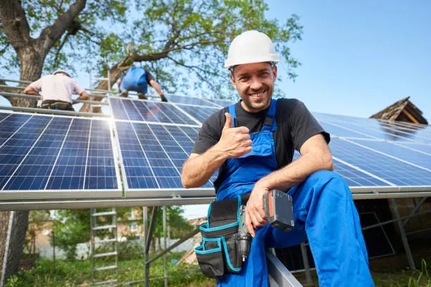 Portrait of smiling technician with electrical screwdriver showing thumb-up in front of unfinished high exterior solar panel photo voltaic system with team of workers on high platform.