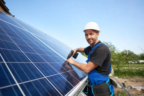Smiling technician mounting solar panel to metal platform using electrical screwdriver on blue sky copy space background. Stand-alone solar system installation, efficiency concept.