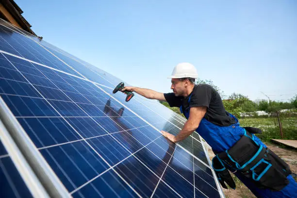 Profile of technician installing solar panel using electrical screwdriver on blue sky copy space background. Stand-alone exterior solar system installation, efficiency and professionalism concept.