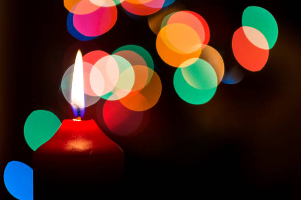 A close-up of a glowing candle inside a decorative lantern adorned with star patterns, creating a warm and magical Christmas ambiance. A blurred ceramic house in the background enhances the cozy holiday setting.