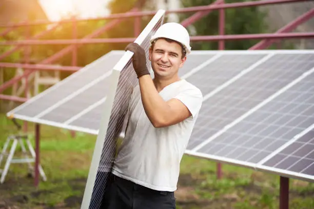 Smiling young worker in protective helmet carrying big shiny solar photo voltaic panel on exterior metal platform background on sunny summer warm day. Renewable ecological green energy concept.