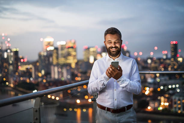 A businessman with smartphone standing against night London view panorama. A portrait of businessman with smartphone standing against night London rooftop view panorama. Copy space. mobile phone lights up stock pictures, royalty-free photos & images