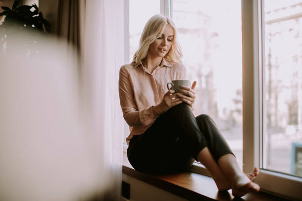 istock Blonde young woman drinking coffee by window 1036740186