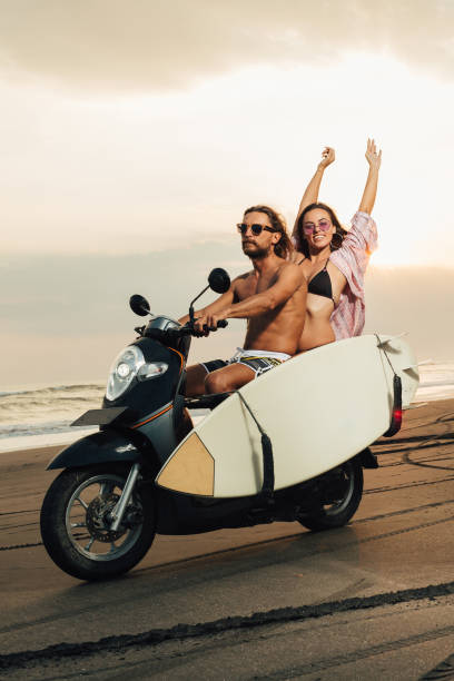 istock boyfriend riding scooter with surfboard on beach, girlfriend sitting with raised hands 1036749420