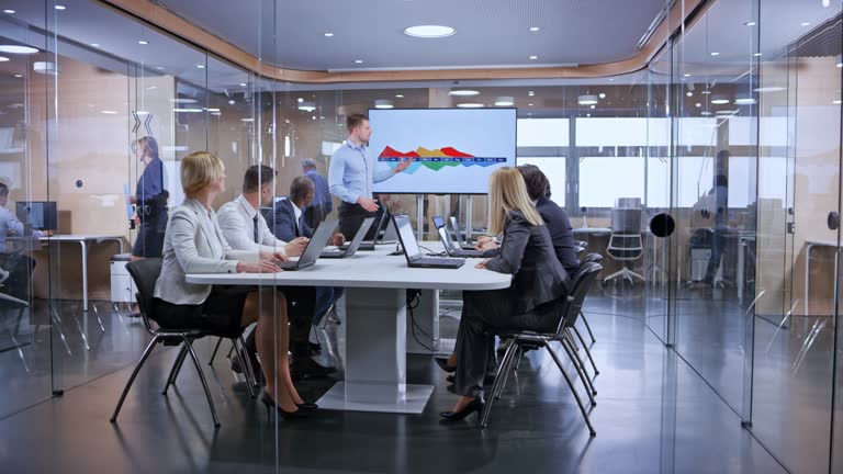 Wide dolly shot of business people sitting in the glass conference room and are attentively listening to the presentation held by their young Caucasian male colleague. Shot in Slovenia.