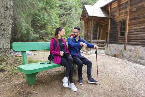 Young couple sitting on a park bench in front of a wooden cabin. Both Caucasian people.