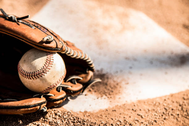 Spring and summer baseball season is here. Wooden bat, glove, and weathered ball lying on home plate in late afternoon sun. Dugout in background. No people. Great background image.
