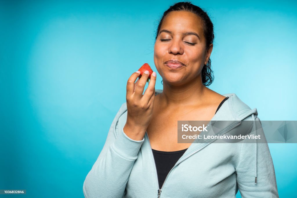 Beautiful young mixed race woman in casual clothing eating and enjoying a fresh strawberry with a blue background Eating Stock Photo