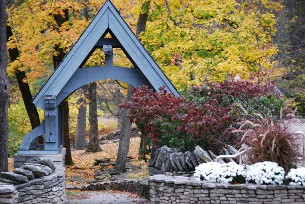 A wooden triangle arch lined by stones surrounded by colorful trees and bushes in the fall at Indiana University in Bloomington, Indiana.