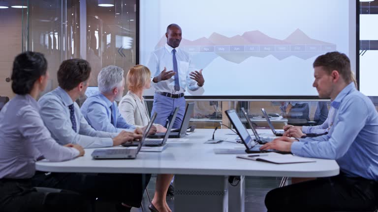Wide dolly shot of an African business man giving a presentation in a glass conference room displaying graphs on the screen for his colleagues. Shot in Slovenia.