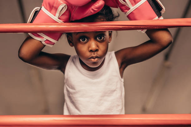 Kid wearing boxing gloves standing near a boxing ring. Close up of a kid boxer standing with hands above the head leaning on boxing ring.