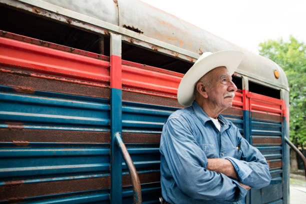 A mexican senior man leaning on a horse box with his arms crossed, thinking and looking away.