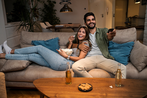 Young Couple Relaxing On Sofa, Chatting Smiling, eatinng popcorn and Changing Channels