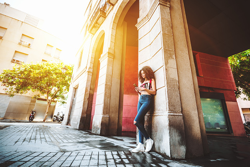 A svelte young charming caucasian female with beautiful curly hair is leaning against the arch column on the street and typing a message to her friend using the smartphone, sunny day in Barcelona
