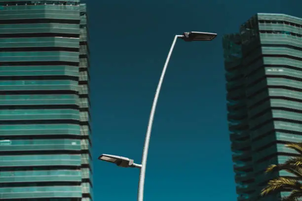 Modern street lantern with two lamps on different height levels in the foreground, huge business office skyscrapers or greenish residential high-rises with glass facades in the defocused background
