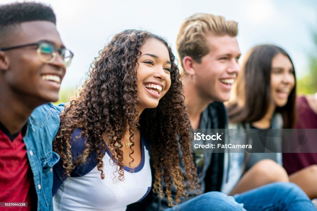 Teenage friends laughing outside A girl of African American descent smiles with laughter while sitting in a row of teens with the arms around each other's shoulders. They are laughing too. Teenager Stock Photo