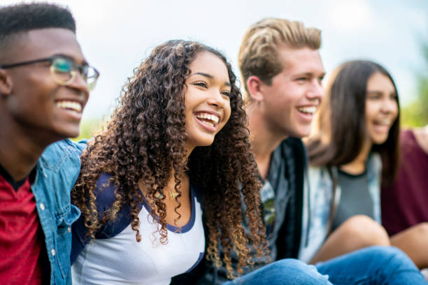 Teenage friends laughing outside A girl of African American descent smiles with laughter while sitting in a row of teens with the arms around each other's shoulders. They are laughing too. social gathering photos stock pictures, royalty-free photos & images