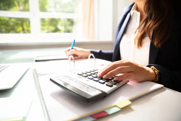 Businesswoman's Hand Calculating Invoice With Calculator In Office