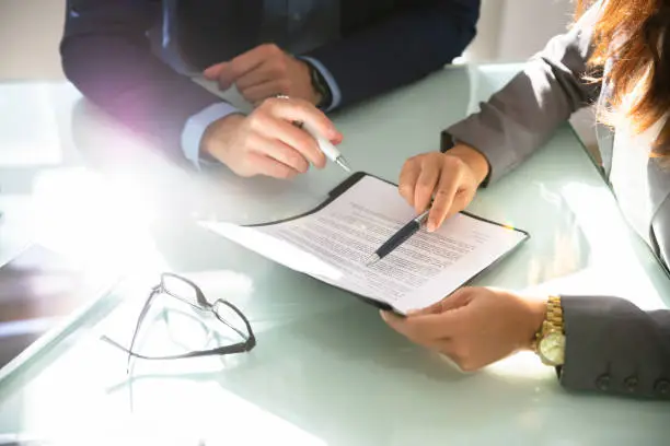 Two Businesspeople Hand Analyzing Document Over Glass Desk