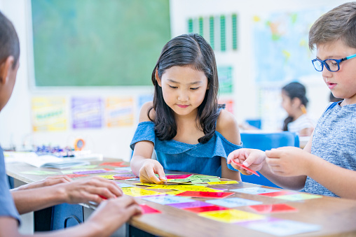 An asian girl and two boys gather around a desk in the classroom and focus on the cards laid out in front of them.